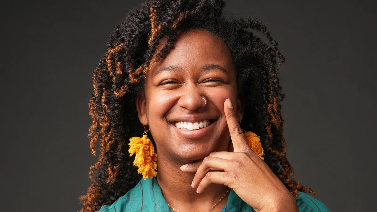 Woman in blue shirt with yellow dangling earrings, smiling at camera with hand on face